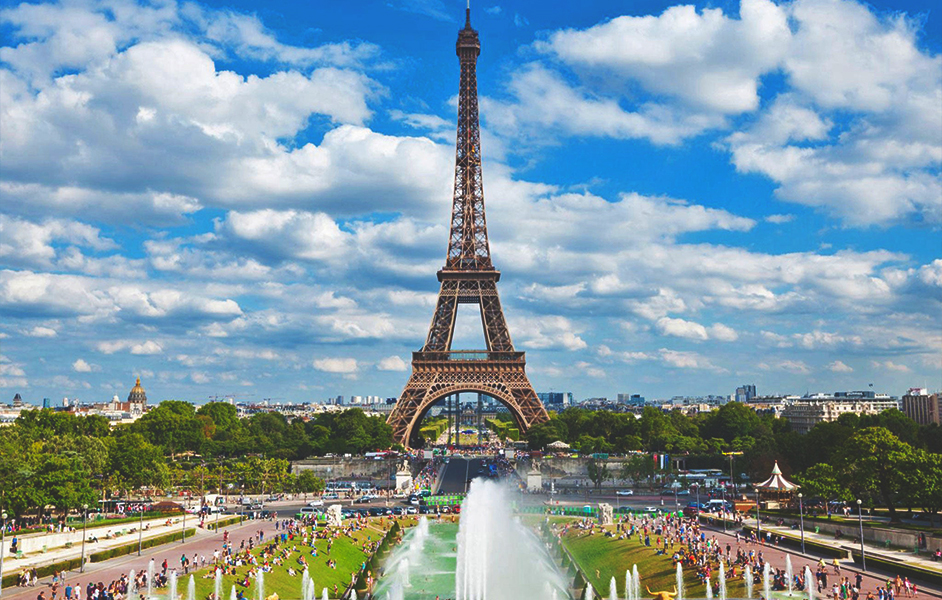 A view of the Eiffel Tower from the Champ de Mars, showcasing the iconic iron lattice structure against a clear blue sky.