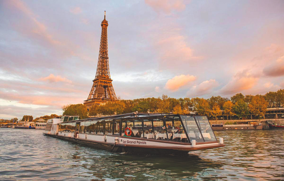 A sightseeing boat cruising on the Seine River with landmarks like the Eiffel Tower and Notre Dame visible in the background.